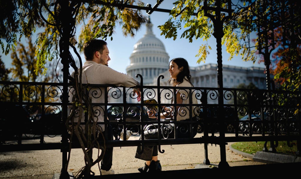 Two people talk on a bench with the U.S. Capitol in the background between them.