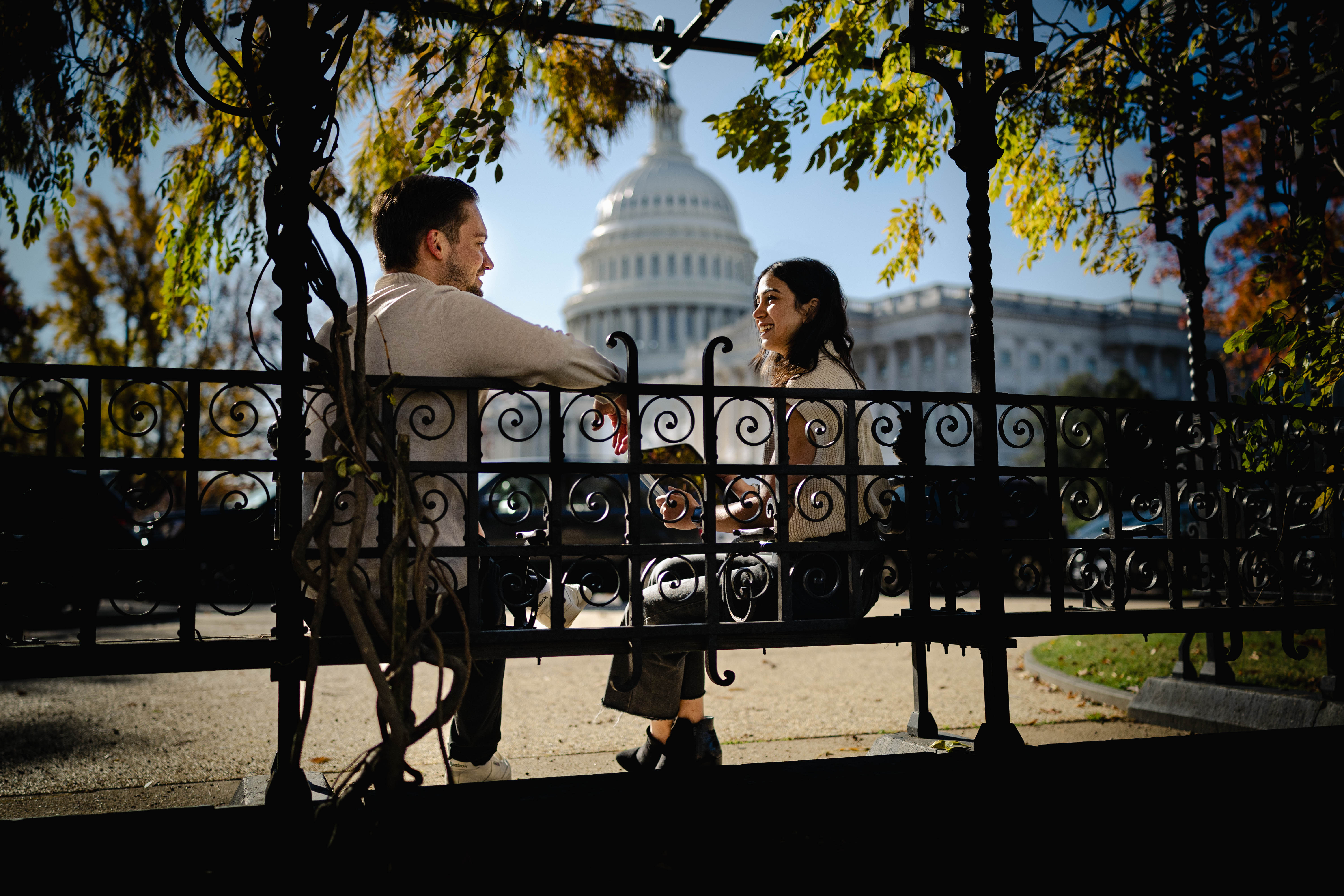 Two people talk on a bench with the U.S. Capitol in the background between them.